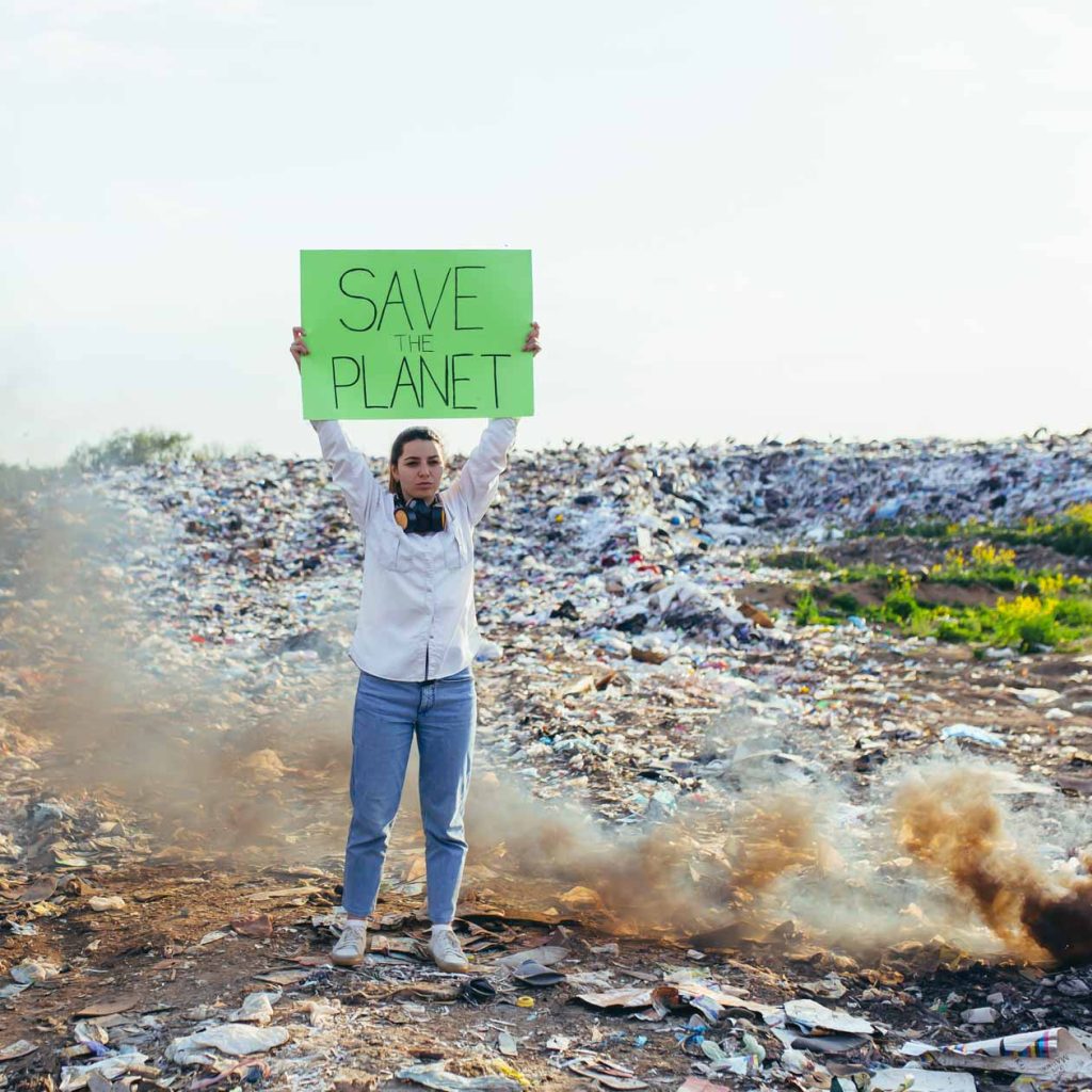 Ein Frau steht auf einer Müllhalde und hält ein Plakat mit Save the planet hoch