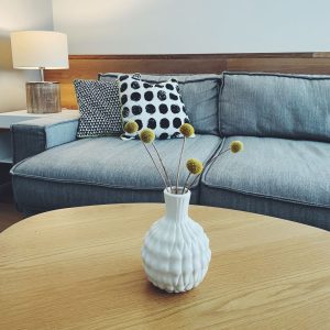 Weiße Vase auf einem Holztisch mit gelben Blumen dahinter eine graue Couch mit getupftem Polster